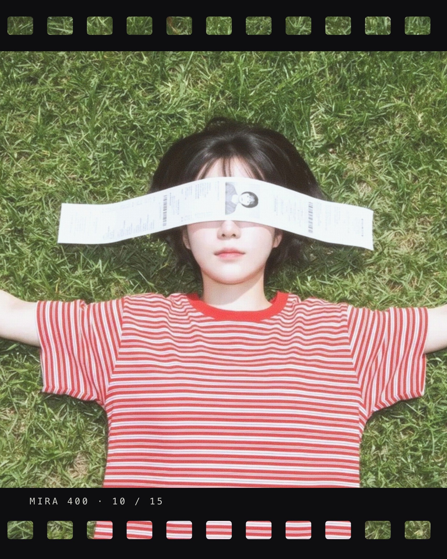 A photograph filling the slide, with a black film strip lying across the top and the bottom and sprocket holes punched through it so the picture shows in them. Overhead on grass, someone in a red and white striped shirt lying with their arms out and a paper strip across their eyes. The label MIRA 400 · 10 / 15 is printed on the strip itself.