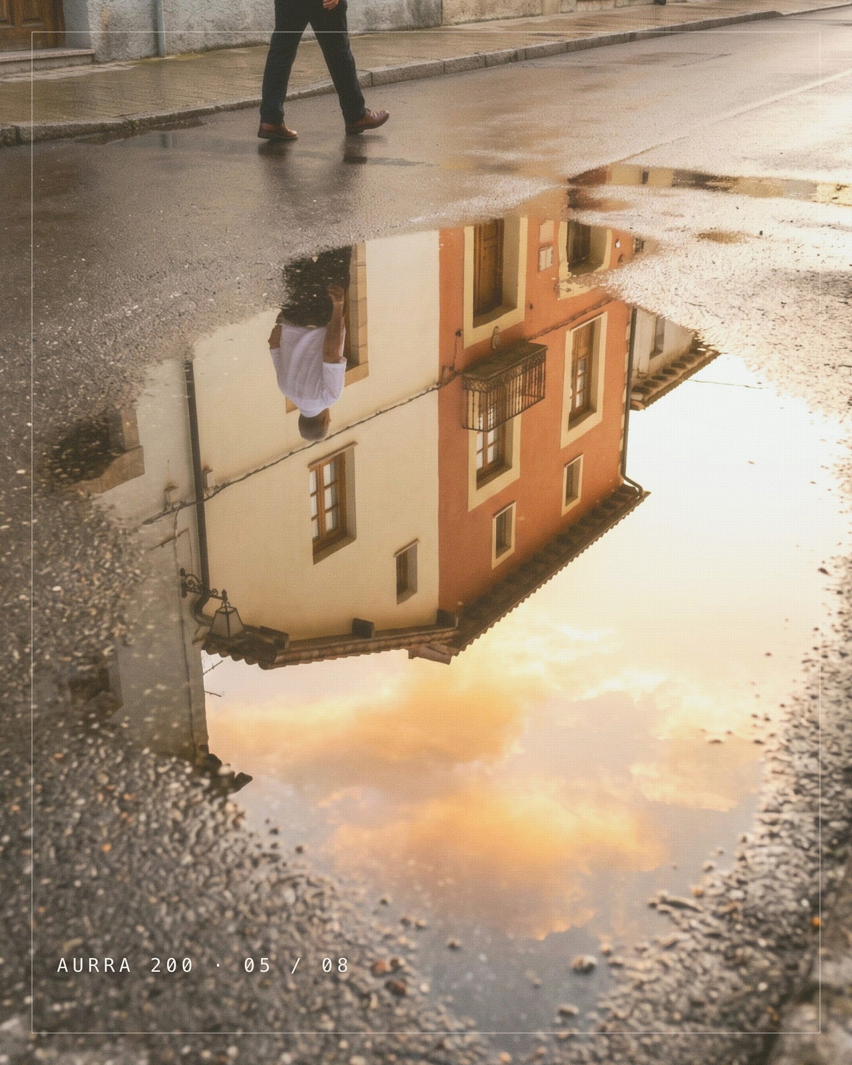 A photograph filling the slide inside a thin paper coloured line, the way a print sits inside its border. A rain puddle on a wet street reflects a cream and orange house and a warm sky, with a man's legs walking past along the top edge. AURRA 200 · 05 / 08 is in mono type at the foot.