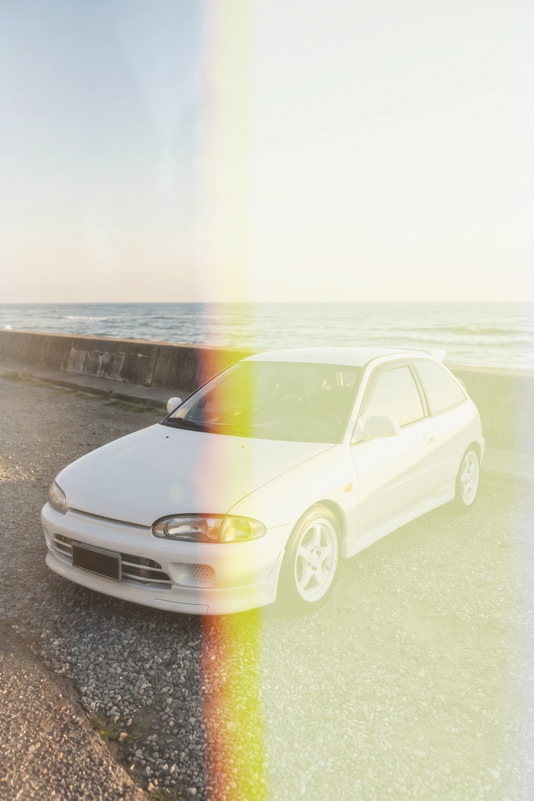 A white hatchback parked on a sea wall, with a band of orange and green burning down the middle of the frame. This is the kind of leak the lab draws, not something the camera did.