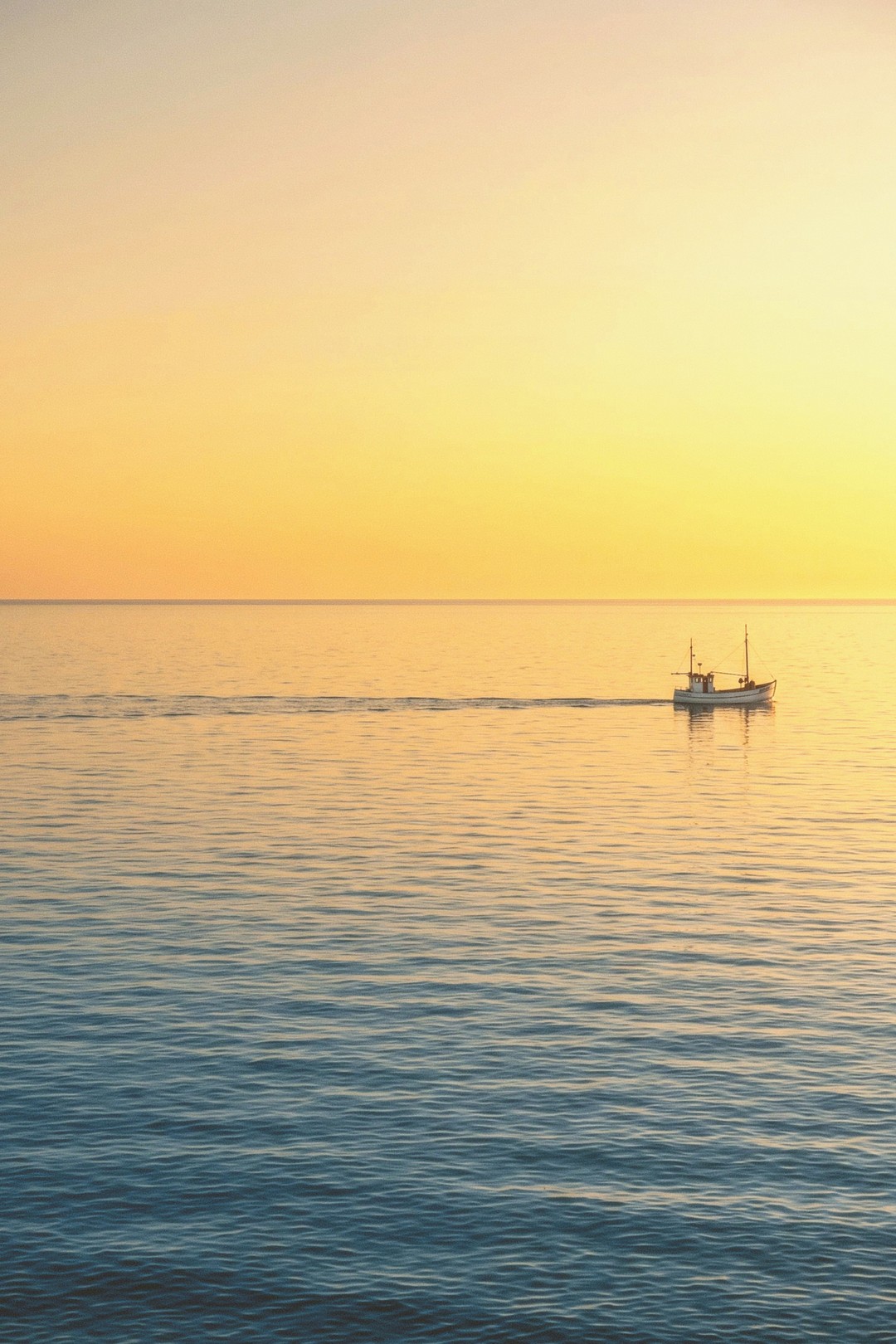 A small fishing boat crossing flat water at sunset, its wake running back across the frame, the sky going from orange at the left to yellow at the right.