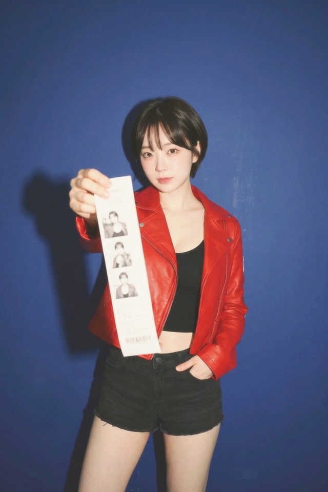 A woman in a red leather jacket against a deep blue wall, holding a paper photo strip out towards the camera. Her shadow is thrown up and to the left, close and hard edged. Mira 400.