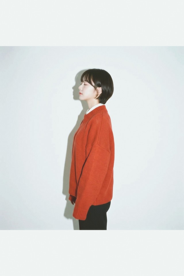 A profile against a pale wall: rust red cardigan, black trousers, looking left. The shadow behind her is soft and barely darker than the wall. Mira 400.